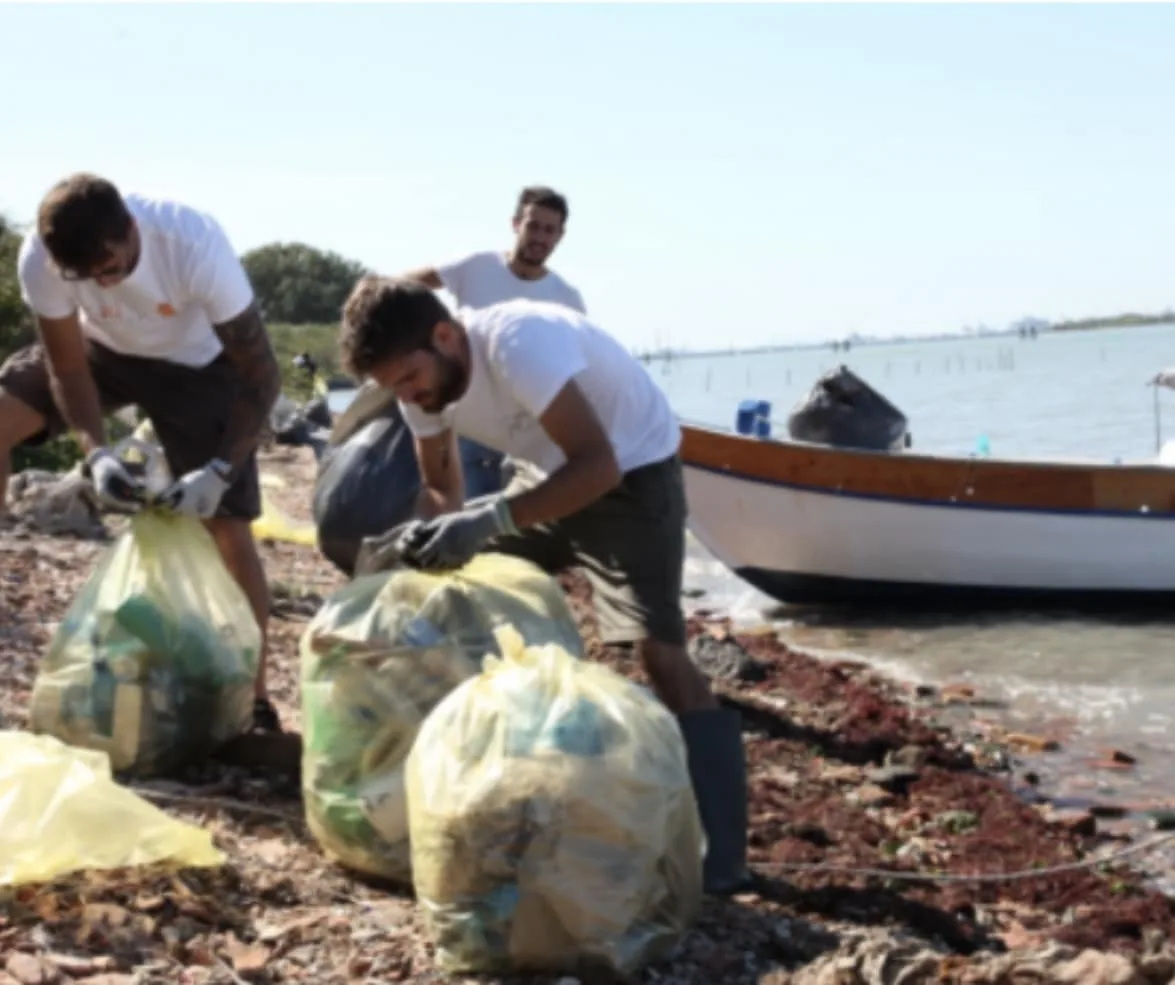 Voyage scolaire durable à Venise : jeunes bénévoles vénitiens en gants remplissant des sacs de déchets sur une rive de la lagune, une barque à l'arrière-plan